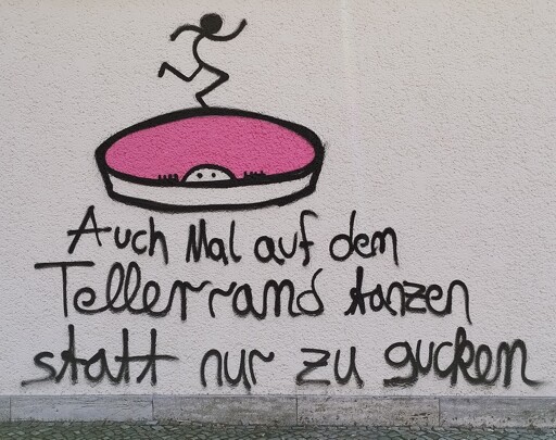 Ein Graffito an einer Wand, mit einer stilisierten Zeichnung eines Tellers, einem Gesicht, das über den Rand lugt, und einer Figur die auf dem Rand des Tellers tanzt, dazu der Text:" Auch mal auf dem Tellerrand tanzen, statt nur zu gucken"